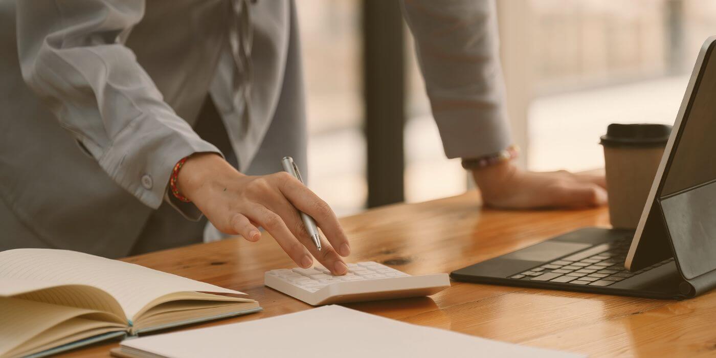 A person in a business suit uses a calculator on a wooden desk, with a notebook and a laptop nearby, analyzing the cost of potential incidents.