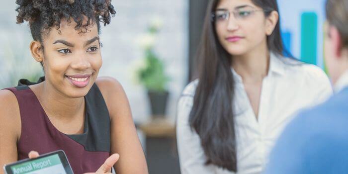 Two women sitting on the same side of a table; one holding a tablet and explaining security risk management to a blurred man on the opposite side of the table