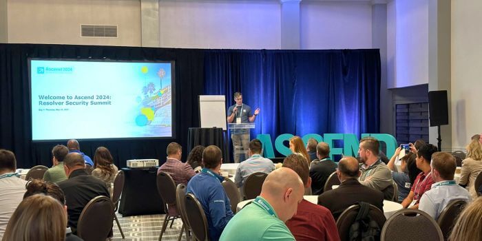A speaker addressing the audience at ascend 2024, with a welcome slide on the screen and attendees seated in a conference room.