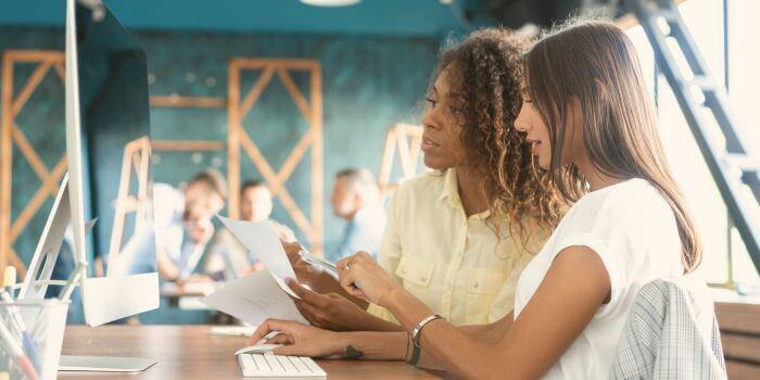 Two women sitting in an office with paperwork in front of them pointing and looking at a computer monitor representing project risk management