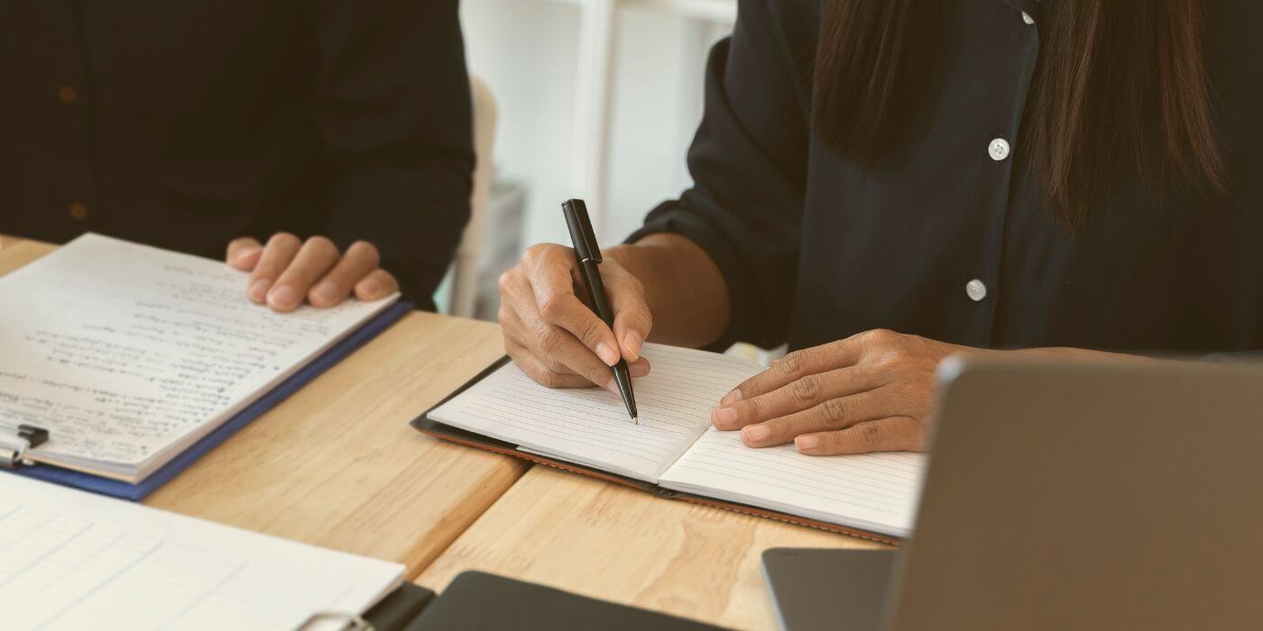 Person taking notes during a meeting with physical security incident reporting software open on a laptop nearby, illustrating organized documentation and software-assisted tracking.