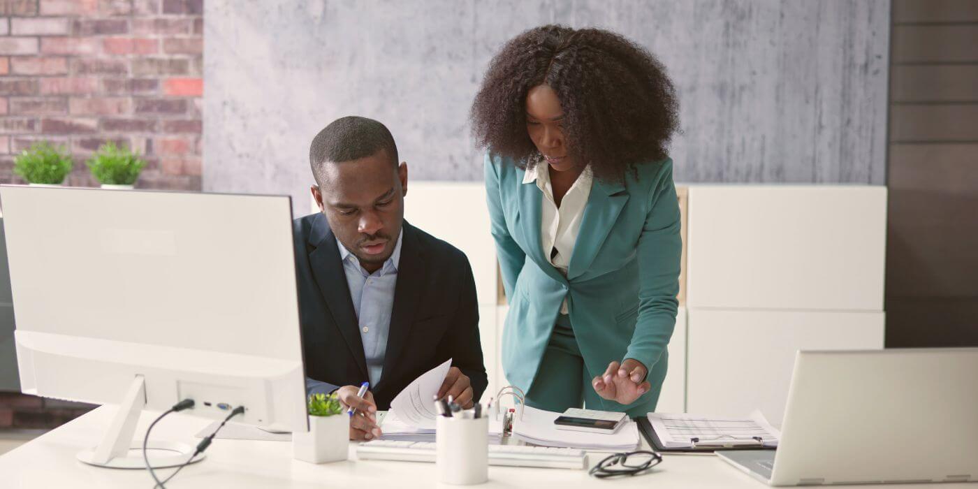 Colleagues collaborate on regulatory change management software, studying documents beside a desktop monitor in a bright, modern office.