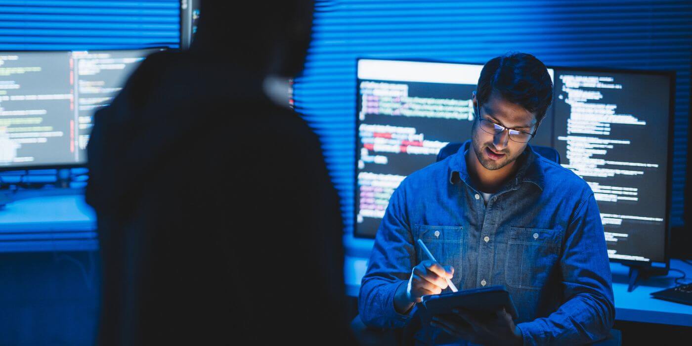 Corporate security investigator reviewing an incident report submission on a digital tablet in a dark control room, with multiple monitors displaying data in the background. Demonstrates ai-powered incident management in action.