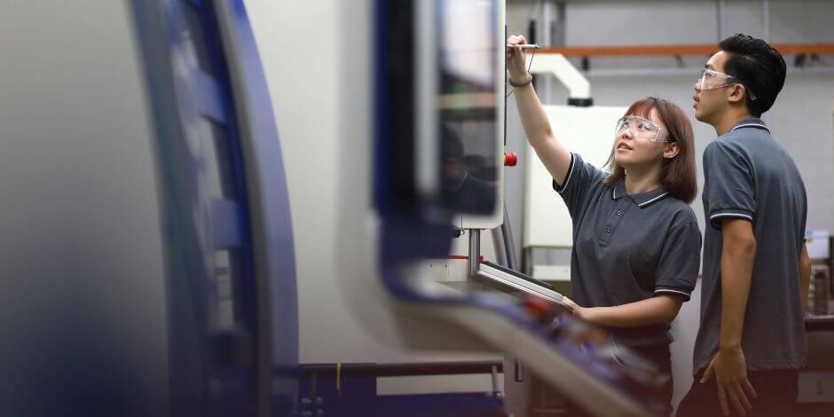 Two people wearing safety glasses and grey collared shirts review information on a control panel in an industrial setting. The image supports content related to speak-up culture and compliance program initiatives, showing how companies like jabil use resolver to strengthen ethical reporting and workplace accountability.