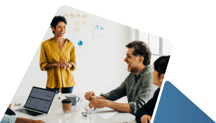 A candid shot of a diverse professional team in a bright office. A woman in a yellow shirt stands and laughs while leading a discussion at a conference table. Intended for testimonial sections.