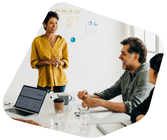 A candid shot of a diverse professional team in a bright office. A woman in a yellow shirt stands and laughs while leading a discussion at a conference table. Intended for testimonial sections.