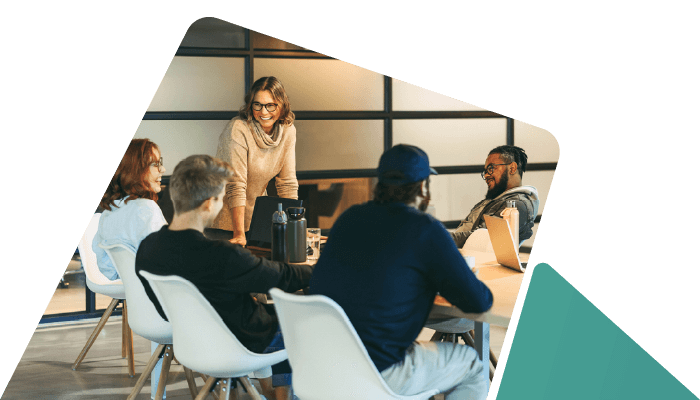 A professional team engaged in a meeting. A woman stands at the head of the table leading a discussion with colleagues seated around a table.
