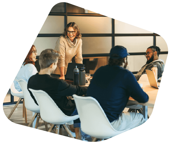 A professional team engaged in a meeting. A woman stands at the head of the table leading a discussion with colleagues seated around a table.