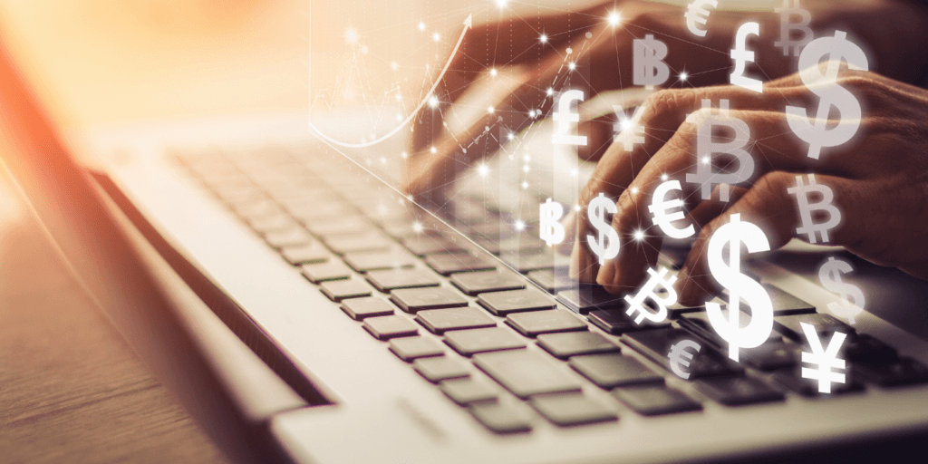 Close-up of a person’s hands typing on a silver laptop keyboard, viewed from the side at desk level. The background is softly blurred with warm lighting. Transparent overlays of currency symbols such as dollar, euro, pound, and yen float above the keyboard alongside thin line graphs and data points, suggesting financial institution risk tied to digital transactions and market activity.